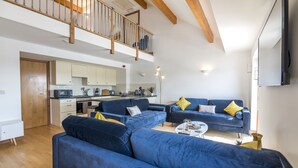 A modern living room with blue sofas, a kitchen in the background, and a wooden staircase.