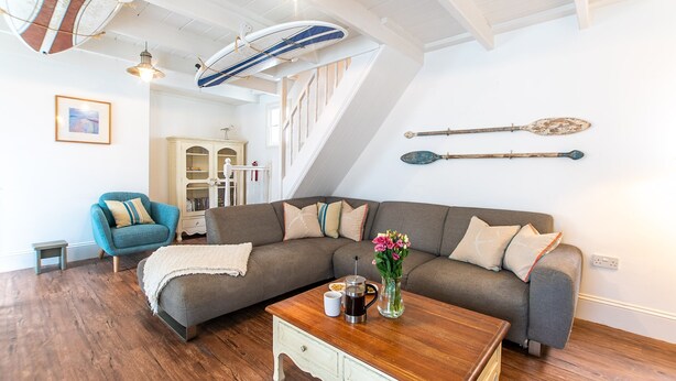 A cozy living room with a large sectional sofa, a coffee table, and a staircase with a white railing. - Stone’s Throw, Central St Ives – Sleeps 7 – Dog Friendly (St Ives)