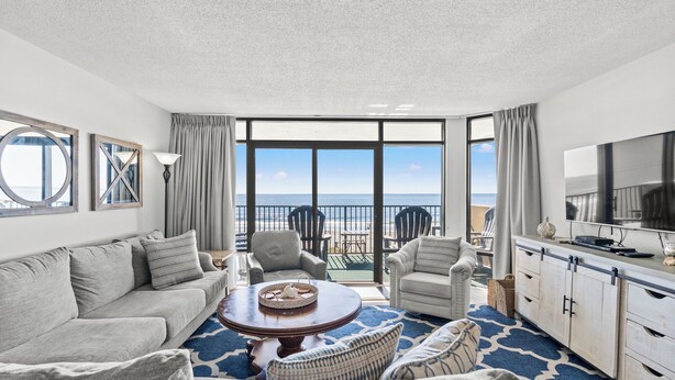 A spacious living room with a large window overlooking the beach, a round coffee table, and a TV on a cabinet. - Verandas 301 - Ocean Front - Luxury 4 Bed/3 Bath Condo (North Myrtle Beach)
