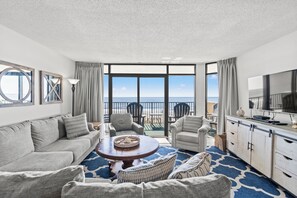Amplia sala de estar con un gran ventanal con vistas a la playa, una mesa de centro redonda y un televisor en un mueble.