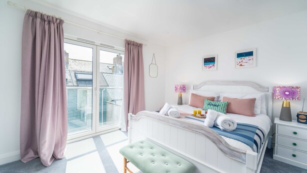 A bedroom with a large window, a bed with pillows and a blanket, a bedside table with a lamp, and a bench.