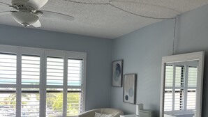Ein Schlafzimmer mit Bett, Sessel, Kommode, Deckenventilator und großen Fenstern.