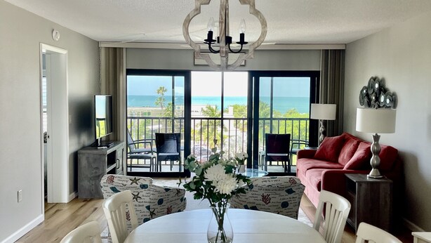 Ein modernes Wohnzimmer mit einem runden Esstisch, weißen Stühlen, einer Chandelüster, einem roten Sofa und Blick auf den Ozean durch große Fenster.