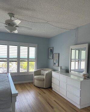 Un dormitorio principal con cama, una silla, cómoda, ventilador de techo y ventanas amplias.