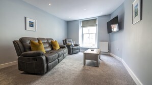 A living room with a leather sofa, two armchairs, a coffee table, and a large window with blinds. - Tipyn O Haul - 1 Bedroom Apartment - Tenby (Tenby)