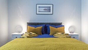 A neatly made bed with a blue and yellow bedspread, two nightstands with round mirrors, and a framed picture on the wall. - Tipyn O Haul - 1 Bedroom Apartment - Tenby (Tenby)