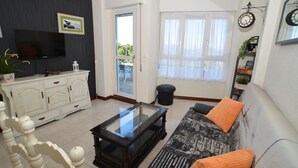 Una sala de estar con televisor, mueble blanco, mesa de centro de vidrio, sofá estampado y vista a través de una puerta de cristal.