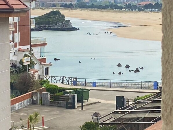 Vista costeira com praia arenosa, afloramento rochoso e um edifício com varandas viradas para o mar.