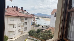 Blick aus einem Fenster auf eine Küstenlandschaft mit einem mehrstöckigen Gebäude, einem Strand und Bergen im Hintergrund.