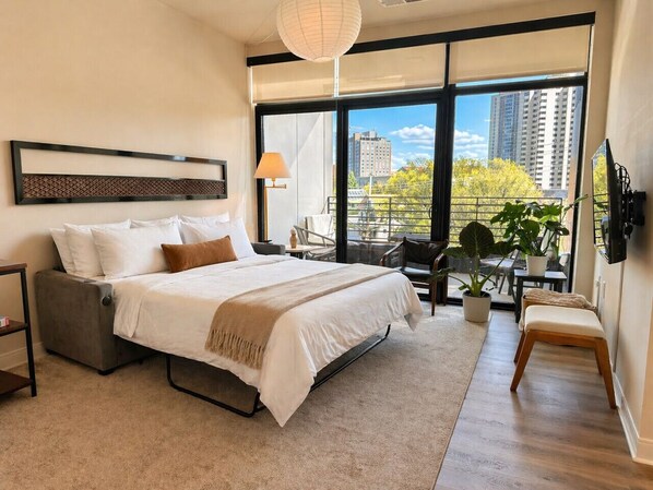 A modern bedroom with a large bed, a sofa, a desk, and a view of the city.