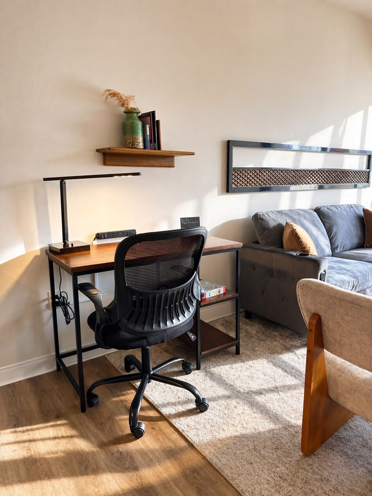 Een moderne woonkamer met een houten bureau, een zwarte bureaustoel, een grijze bank en een houten stoel.