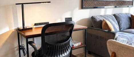 Een moderne woonkamer met een houten bureau, een zwarte bureaustoel, een grijze bank en een houten stoel.