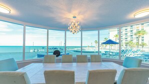 A dining area with a large table and chairs, large windows with a view of the beach and pool area.