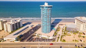 A tall, cylindrical hotel with a blue and white facade, located adjacent to a beach with a parking lot and a road in the foreground.