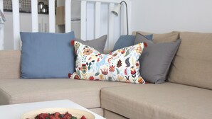 Un sofá color beige con cojines azules y grises, una mesa blanca con un cuenco de fruta y un cojín estampado.