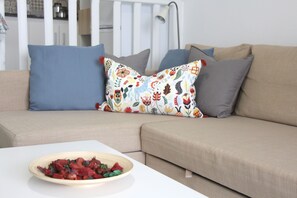 Un divano beige con cuscini blu e grigi, un tavolo bianco con una ciotola di frutta e un cuscino decorato.