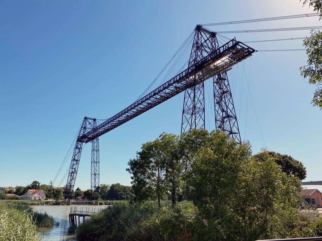 A large metal bridge structure with a truss design, spanning over a body of water with trees and buildings in the background.