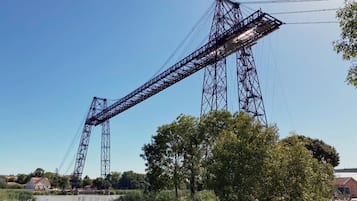 A large metal bridge structure with a truss design, spanning over a body of water with trees and buildings in the background.