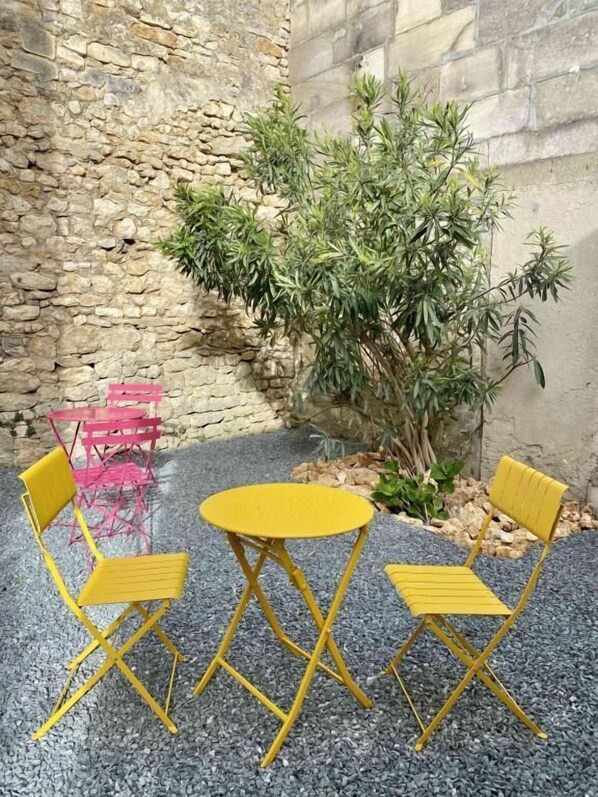 A small outdoor seating area with yellow chairs and a table, surrounded by a stone wall and a small tree.