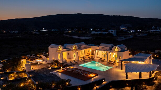A large, white, Mediterranean-style building with a swimming pool, outdoor seating, and a view of a hilly landscape. - Santa Irina Luxury Sunny Lofts 4 (ΣΑΝΤΟΡΙΝΗ)