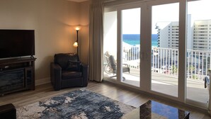 A living room with a TV, a chair, a lamp, and a view of the ocean through large windows.