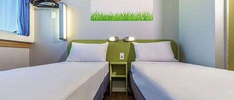 A hotel room with two beds, a nightstand, a wall-mounted lamp, and a window with curtains.