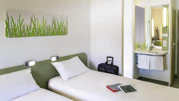 Habitación de hotel con dos camas, una mesita de noche, un escritorio pequeño, un baño con lavamanos y espejo, y un cuadro de pared con motivos verdes.