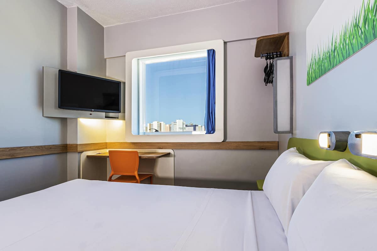 Habitación de hotel con cama, escritorio, silla, televisor montado en la pared, ventana con cortina y un cuadro de hierba en la pared.