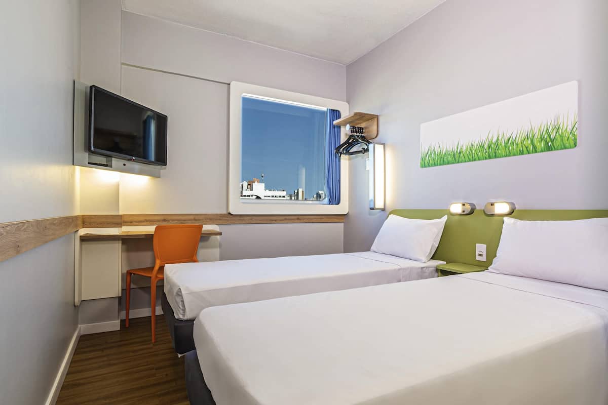 Habitación de hotel con dos camas, un escritorio, una silla, un televisor, una ventana con vista a la ciudad y un cuadro de césped.