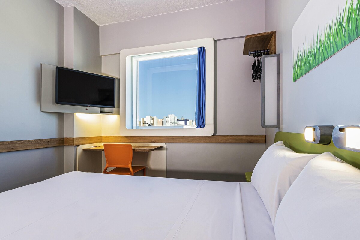 A hotel room with a bed, a desk, a chair, a TV mounted on the wall, a window with a curtain, and a painting of grass on the wall.