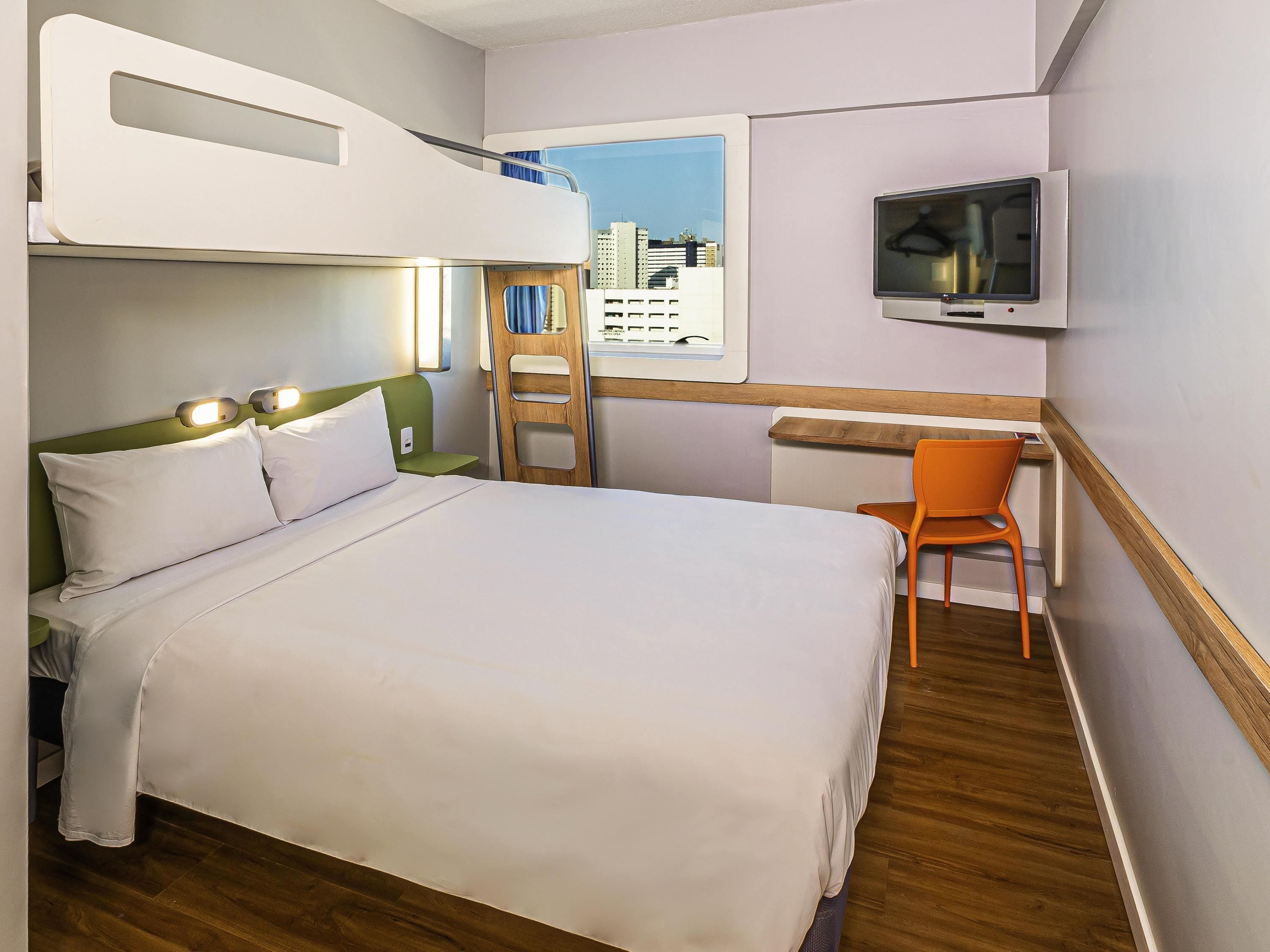 Quarto de hotel com uma cama beliche, uma mesa com cadeira, uma televisão e uma janela com vista para a cidade.