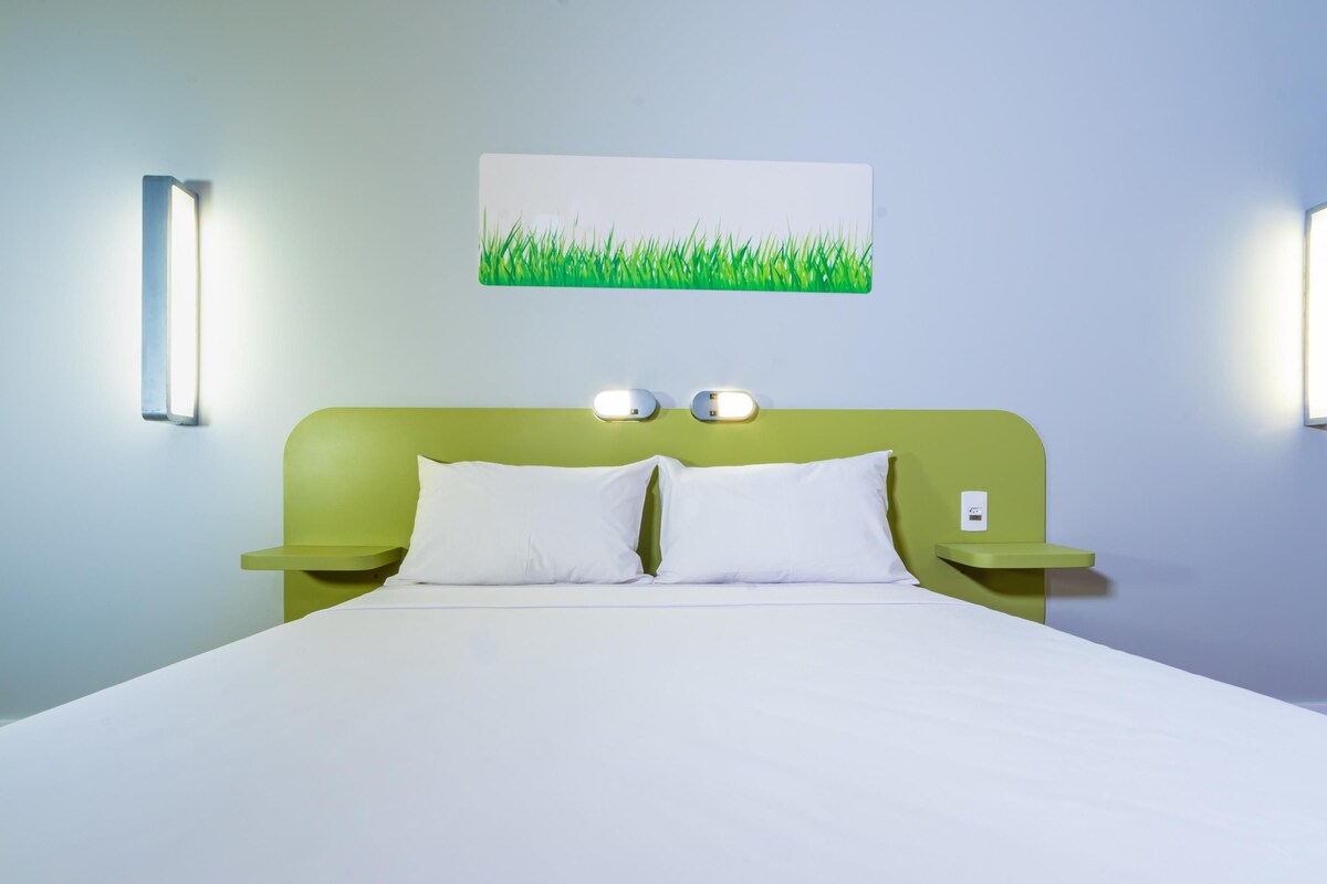 Quarto de hotel com cabeceira verde, roupa de cama branca e duas mesas de cabeceira com luminárias fixadas na parede.