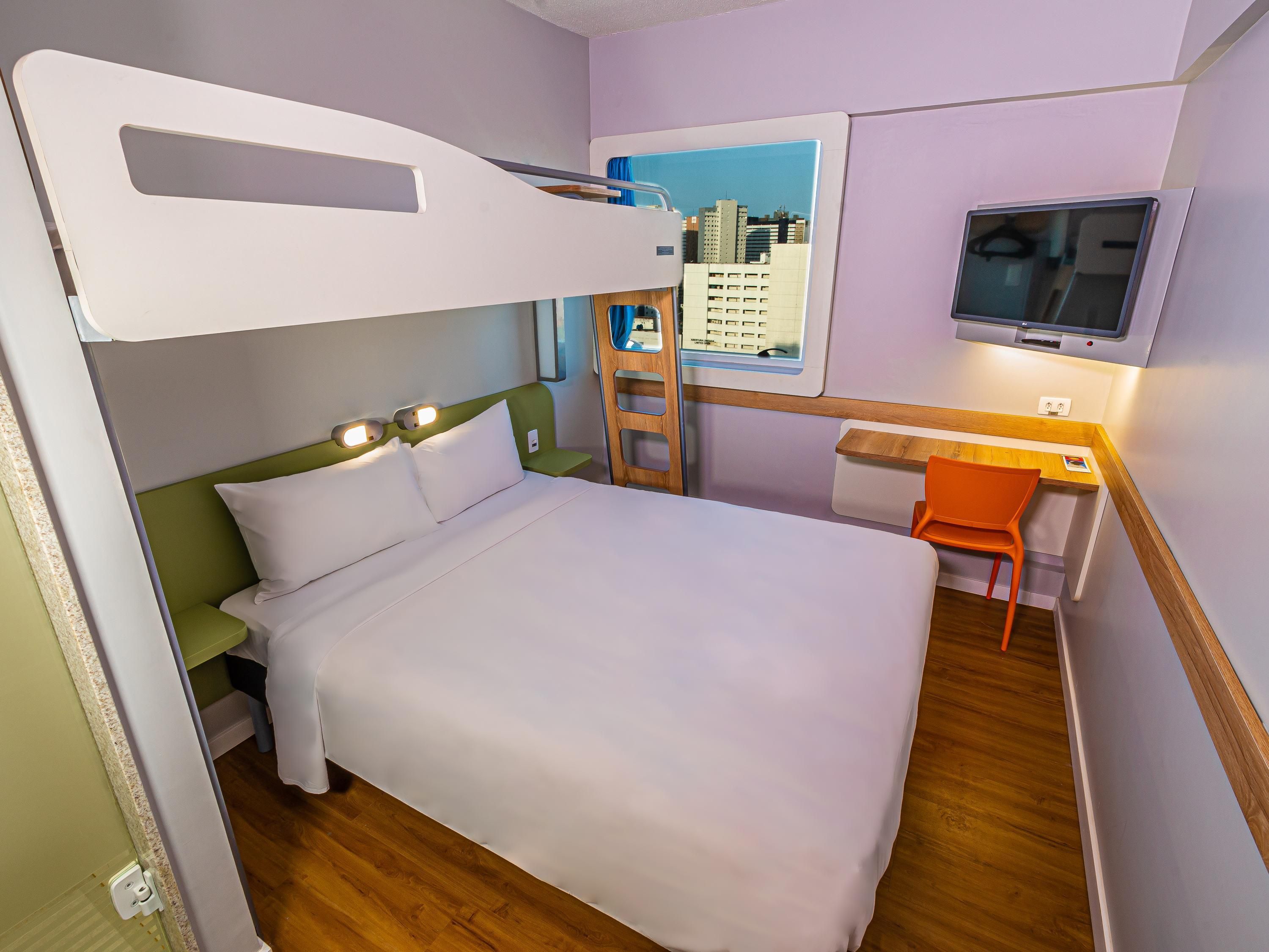 A bunk bed with a desk and chair, a television, and a window with a cityscape view.