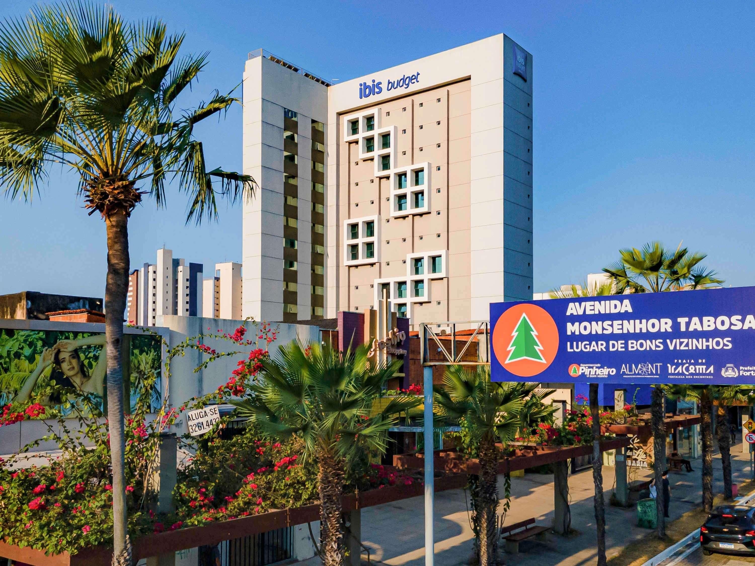 A modern hotel with a prominent 'ibis budget' sign, surrounded by palm trees and a signboard for 'AVENIDA MONSENHOR TABOSA LUGAR DE BONS VIZINHOS.'