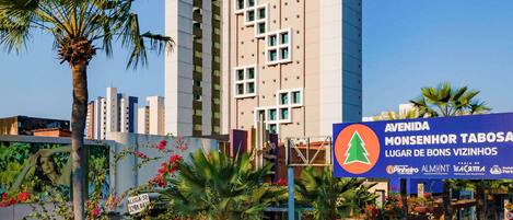 A modern hotel with a prominent 'ibis budget' sign, surrounded by palm trees and a signboard for 'AVENIDA MONSENHOR TABOSA LUGAR DE BONS VIZINHOS.'