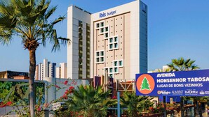 Un hotel moderno con un llamativo letrero de 'ibis budget', rodeado de palmeras y un cartel que indica 'AVENIDA MONSENHOR TABOSA LUGAR DE BONS VIZINHOS'.