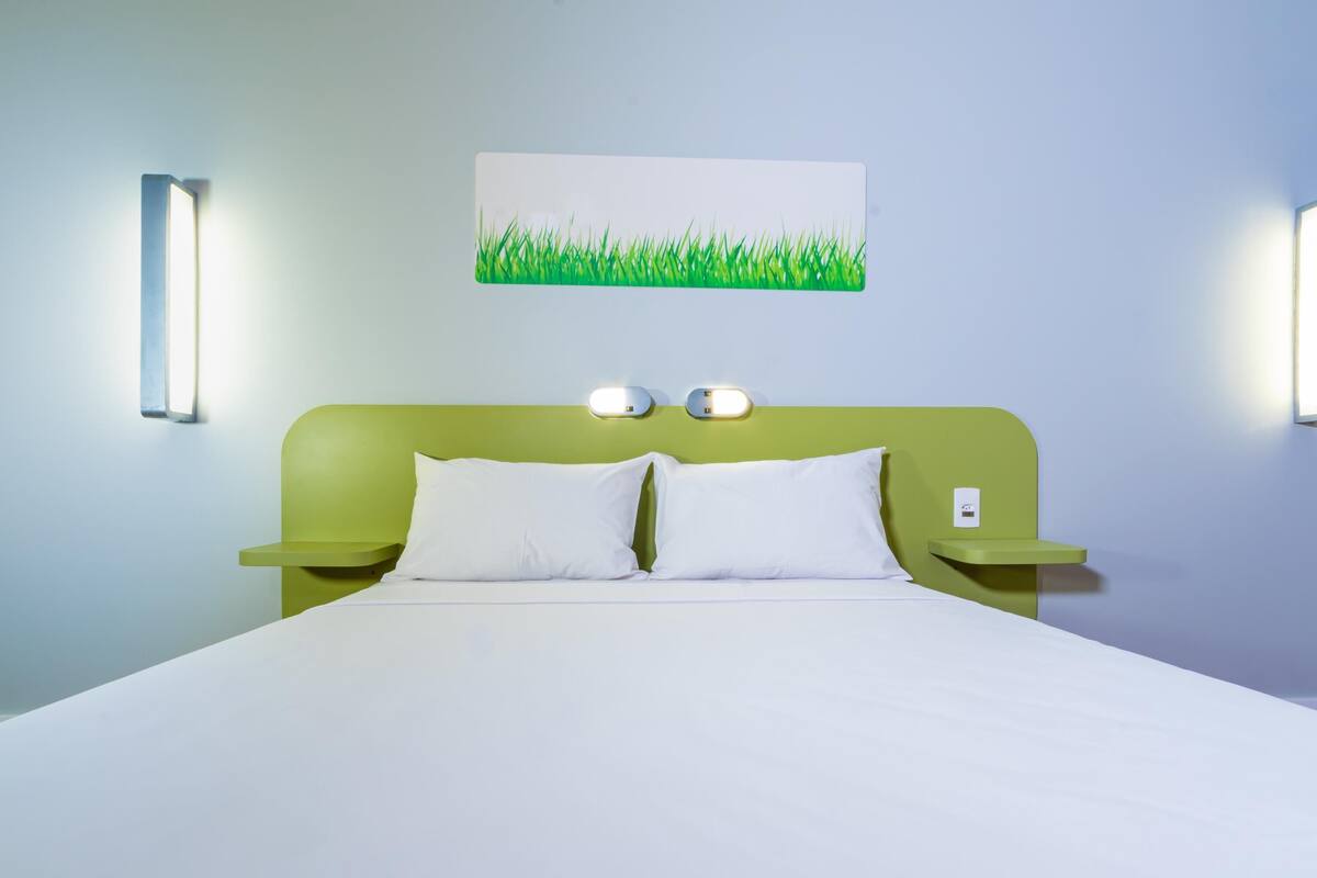 A hotel room with a green headboard, white bedding, and two bedside tables with wall-mounted lamps.