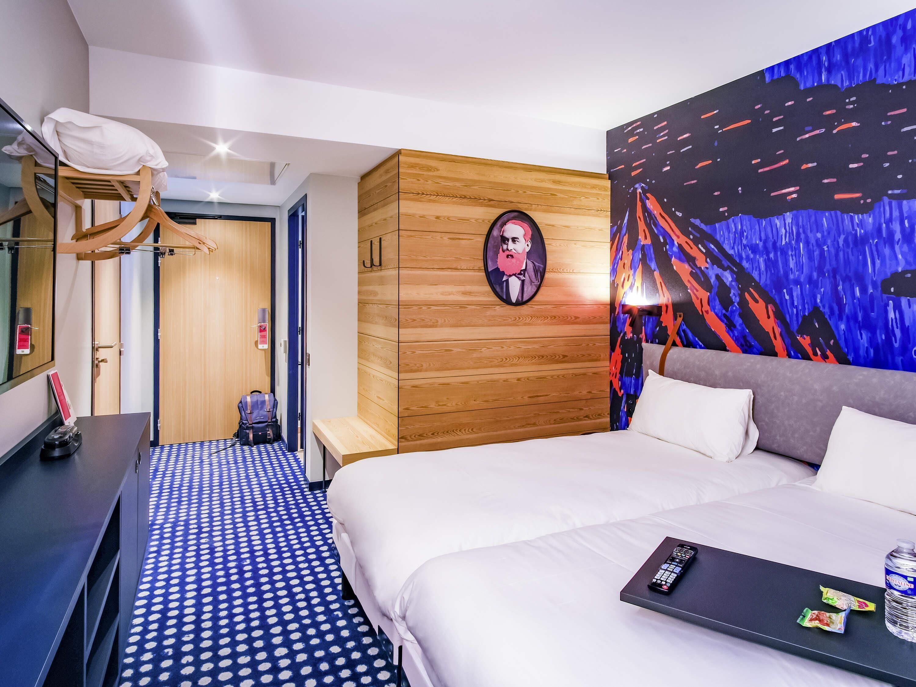 Ein Hotelzimmer mit Bett, Schreibtisch, Stuhl, Kleiderschrank und einer Wand mit einem farbenfrohen abstrakten Gemälde.