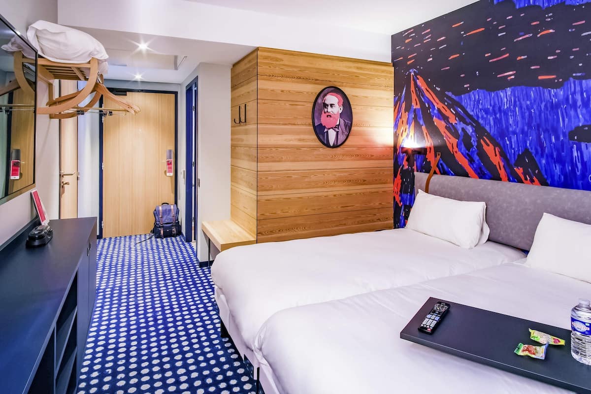 Une chambre d’hôtel comprenant un lit, un bureau, une chaise, une armoire et un mur orné d’un tableau abstrait aux couleurs vives.