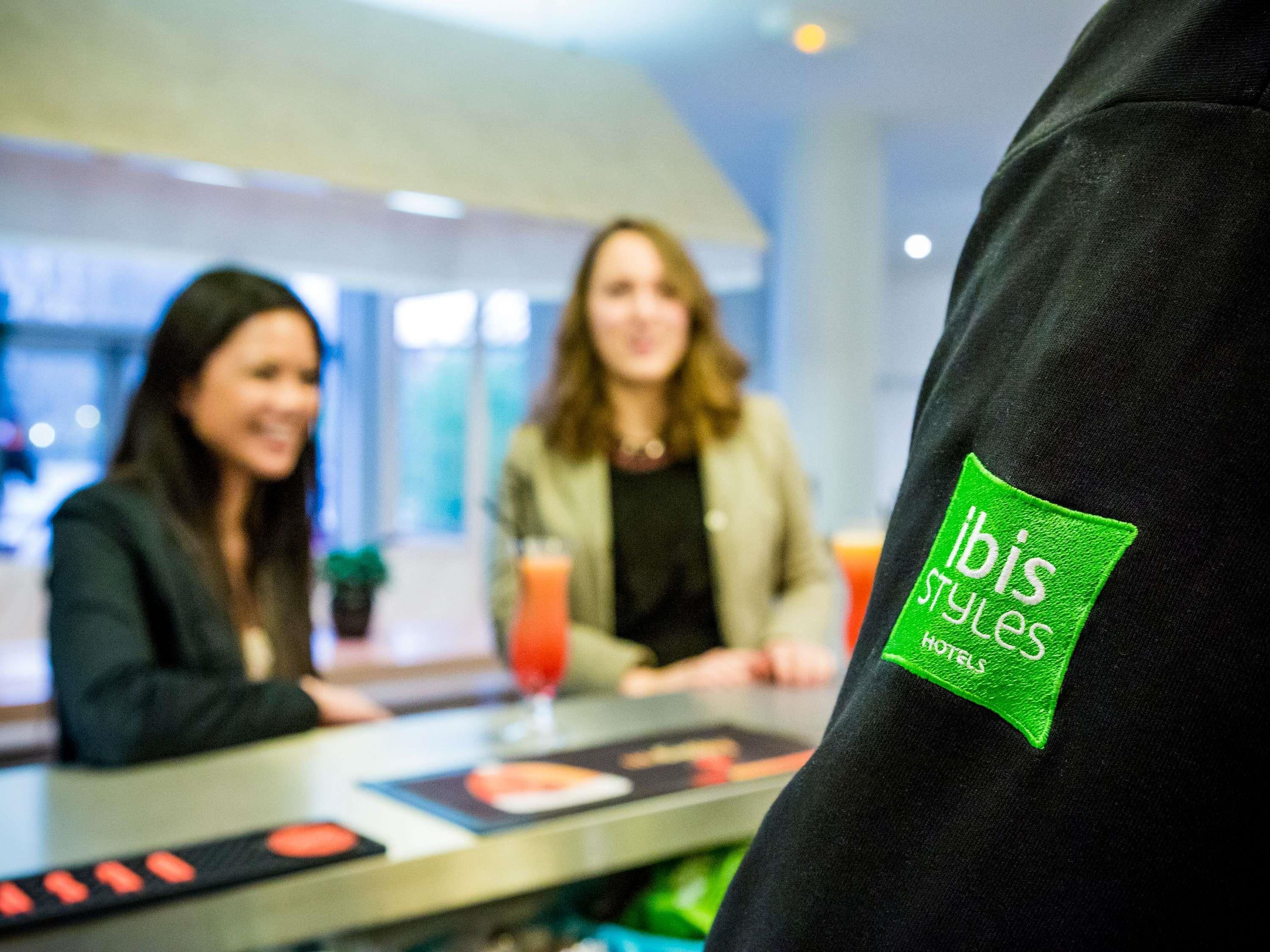 Eine Person trägt eine schwarze Jacke mit einem grünen Etikett, auf dem 'ibis styles hot' steht.