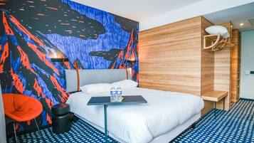 Ett modernt hotellrum med en säng, ett litet bord med två vattenflaskor, en stol och en unik väggmålning.