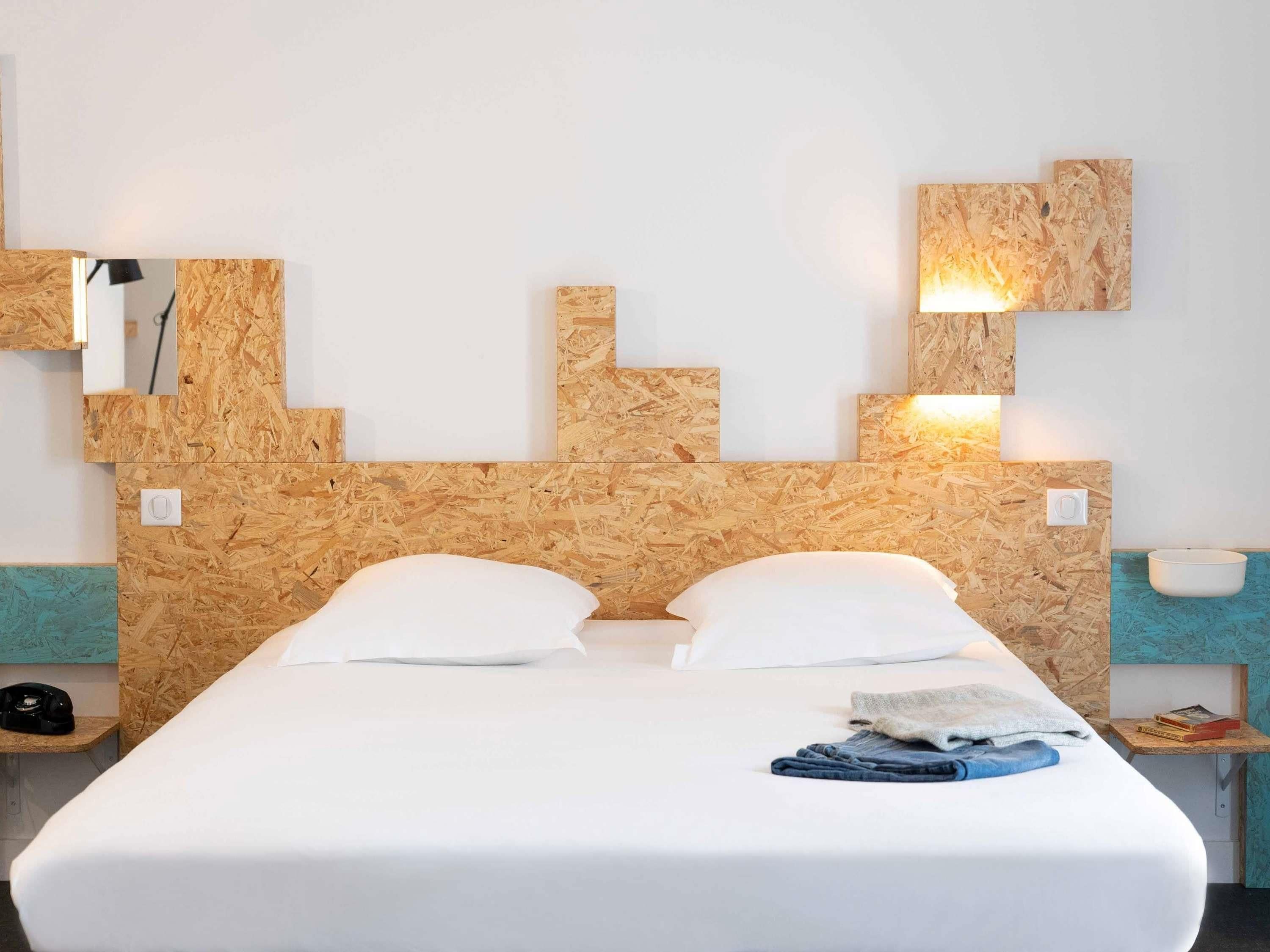 Een hotelkamer met een houten hoofdeinde, twee witte kussens, een netjes opgemaakt bed, een nachtkastje met een lamp en een klein plankje met boeken.