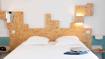 Een hotelkamer met een houten hoofdeinde, twee witte kussens, een netjes opgemaakt bed, een nachtkastje met een lamp en een klein plankje met boeken.