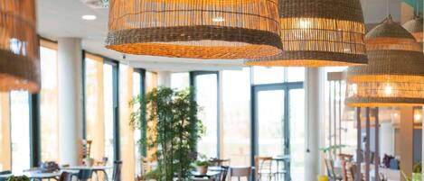 A spacious dining area with long tables, wicker pendant lighting, and large windows providing natural light.