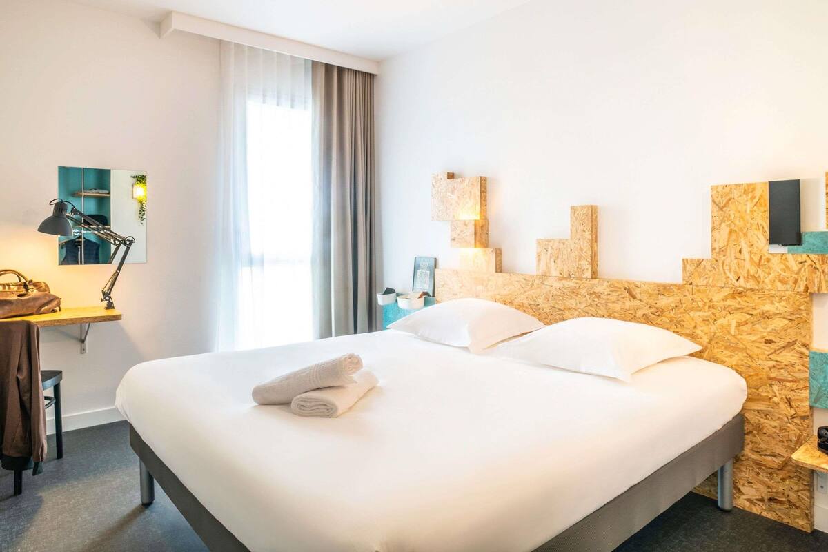 Una habitación de hotel con una cama, un escritorio con lámpara, un espejo y una ventana con cortinas.
