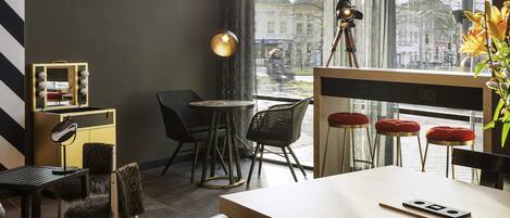 A modern hotel room with a large window, a desk, chairs, a vanity with lights, and a tripod.