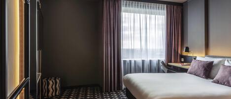 Una habitación de hotel con cama, escritorio con lámpara, una silla y alfombra estampada.