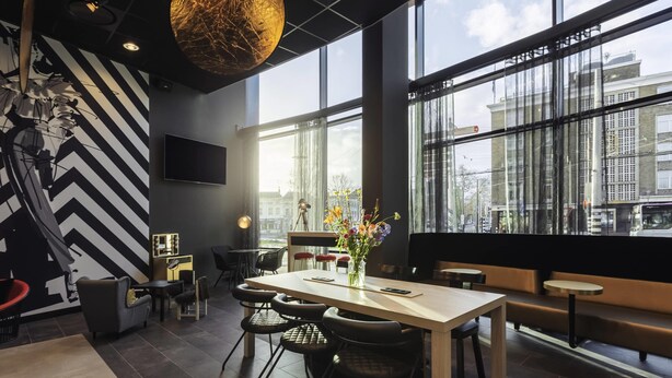 A modern interior with a large window, a dining area with chairs and a table, and a decorative wall with a geometric pattern. - ibis Styles Arnhem Centre (Arnhem)