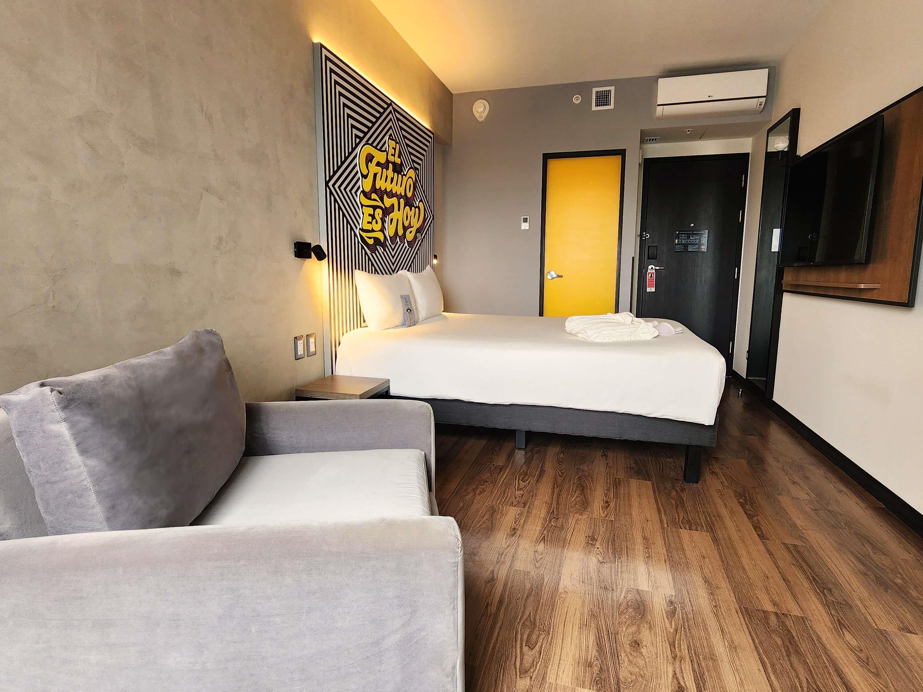A hotel room with a bed, a grey sofa, a wall-mounted TV, and a wooden floor.