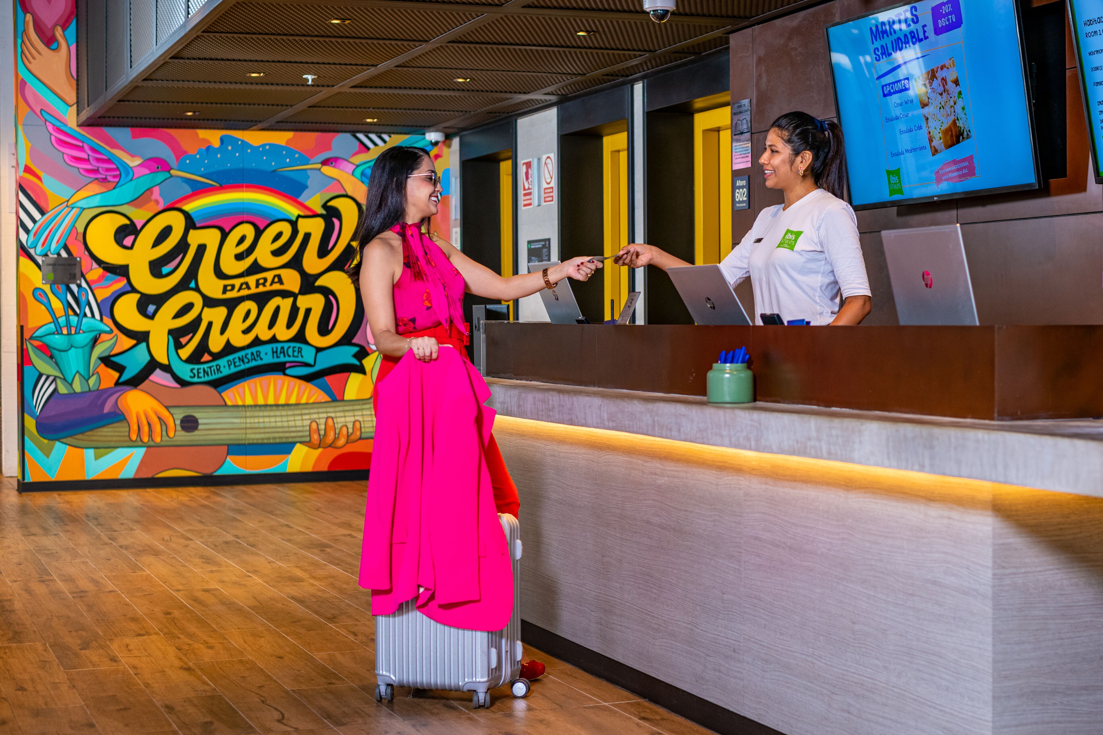 A hotel reception area with a colorful mural, a reception desk, and a luggage cart.